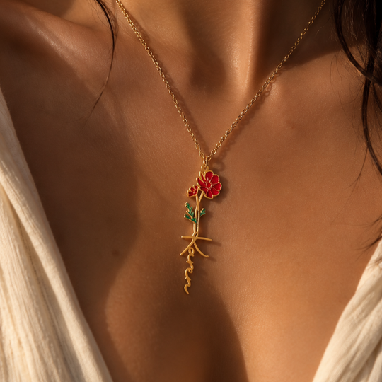 A close-up lifestyle shot of a model wearing a delicate gold necklace with a rose pendant. The pendant features a red rose with green leaves and gold roots, set against warm, sunlit skin.