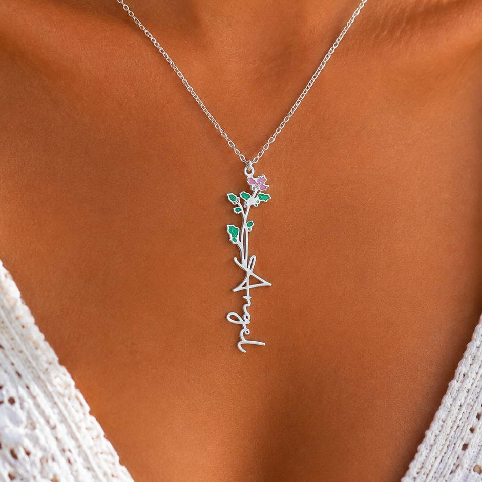 A close-up of a model wearing a delicate silver chain necklace with a script name pendant and small green floral accents.