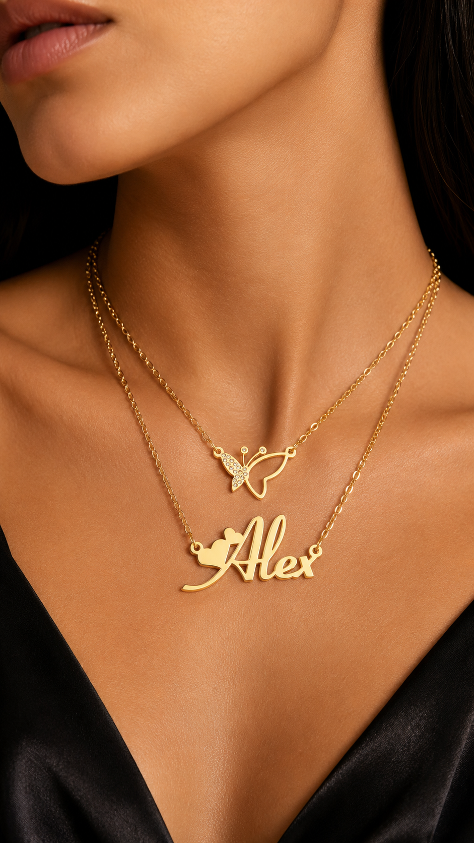 A close-up of a model wearing a layered gold necklace set. The top layer features a delicate butterfly pendant, while the bottom layer has a personalized nameplate pendant spelling 'Alex' with a small heart accent.
