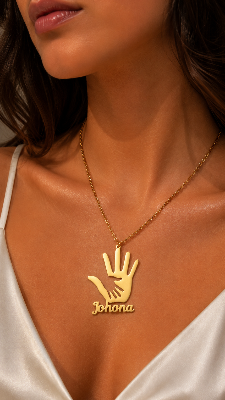 A close-up of a woman wearing a gold necklace with a hand-shaped pendant that has the name 'Johona' engraved below it, paired with a white satin top.