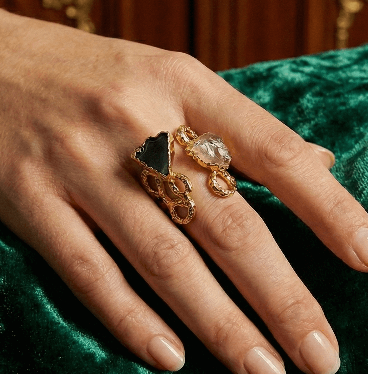 Hand wearing a gold ring with a black stone and clear gemstone on a green velvet surface.