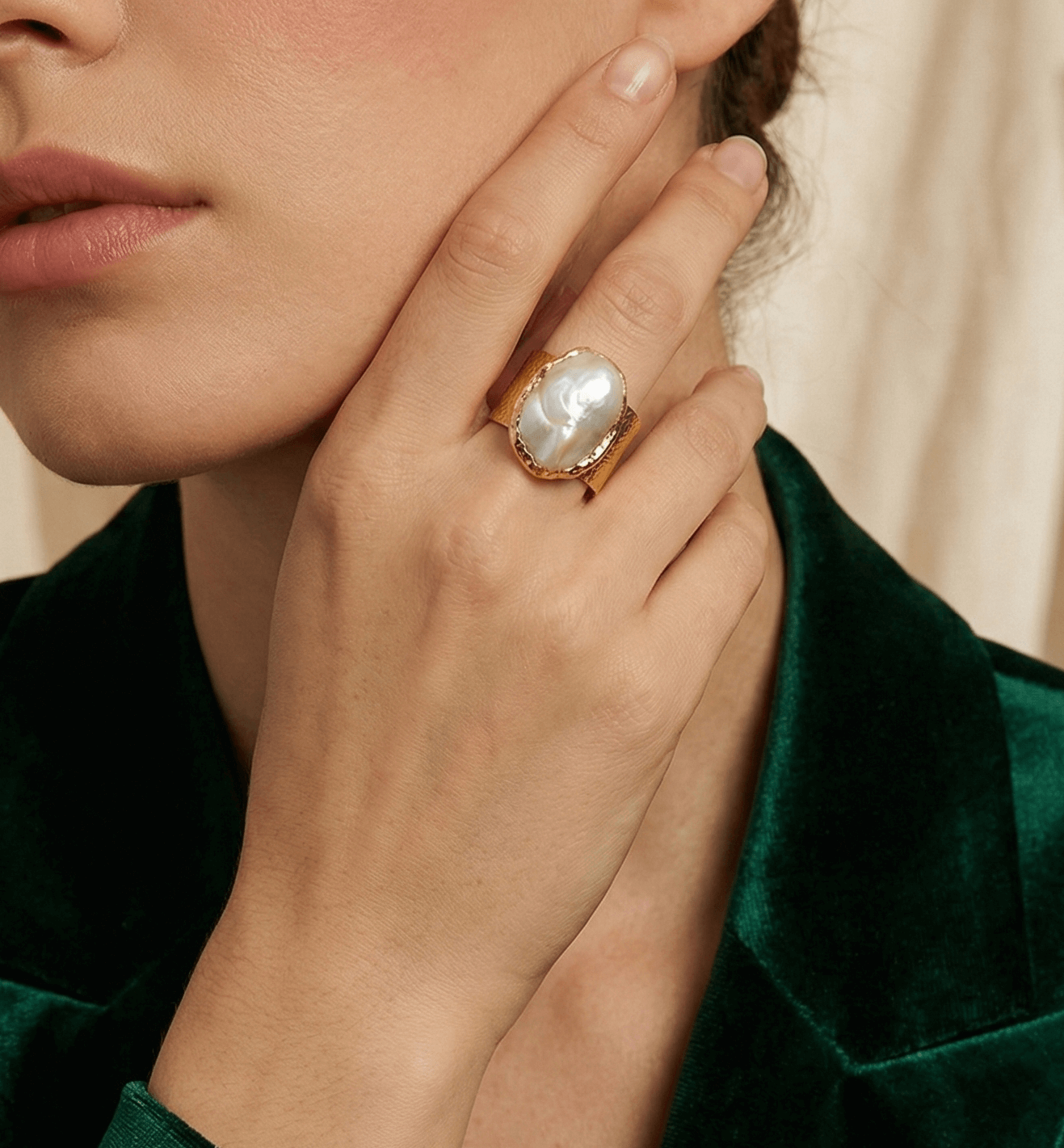 Close-up of a hand wearing a gold ring with a large gemstone, held near the face against a neutral background.