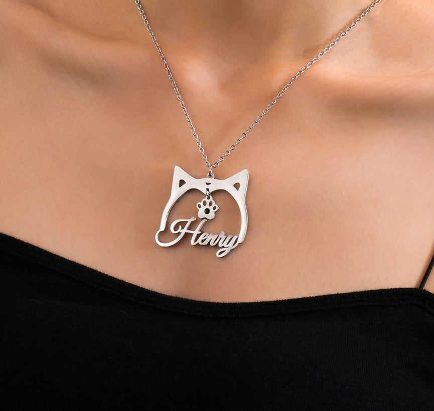 A close-up of a woman wearing a silver cat-shaped pendant necklace with the name 'Hery' engraved on it, paired with a black spaghetti-strap top against a neutral gray background.